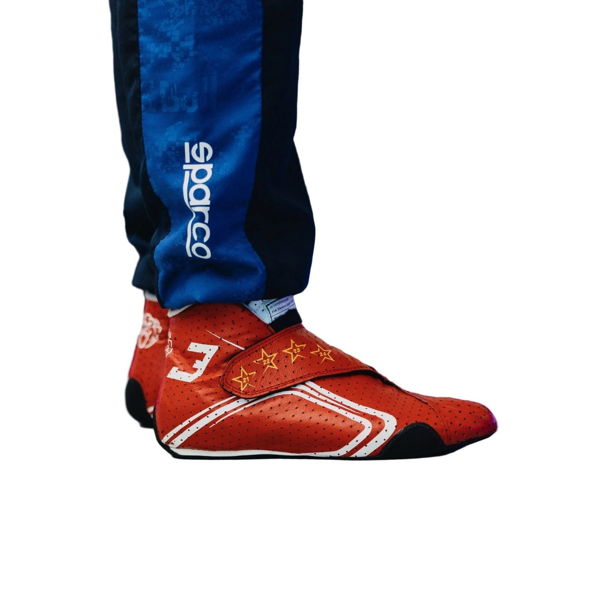 2026 Racing Shoes Replica – Max Verstappen Monaco GP: a close-up of a racing shoe worn with Sparco pants, featuring a red upper with white and yellow graphics, black sole, and blue pants with a white Sparco logo.
