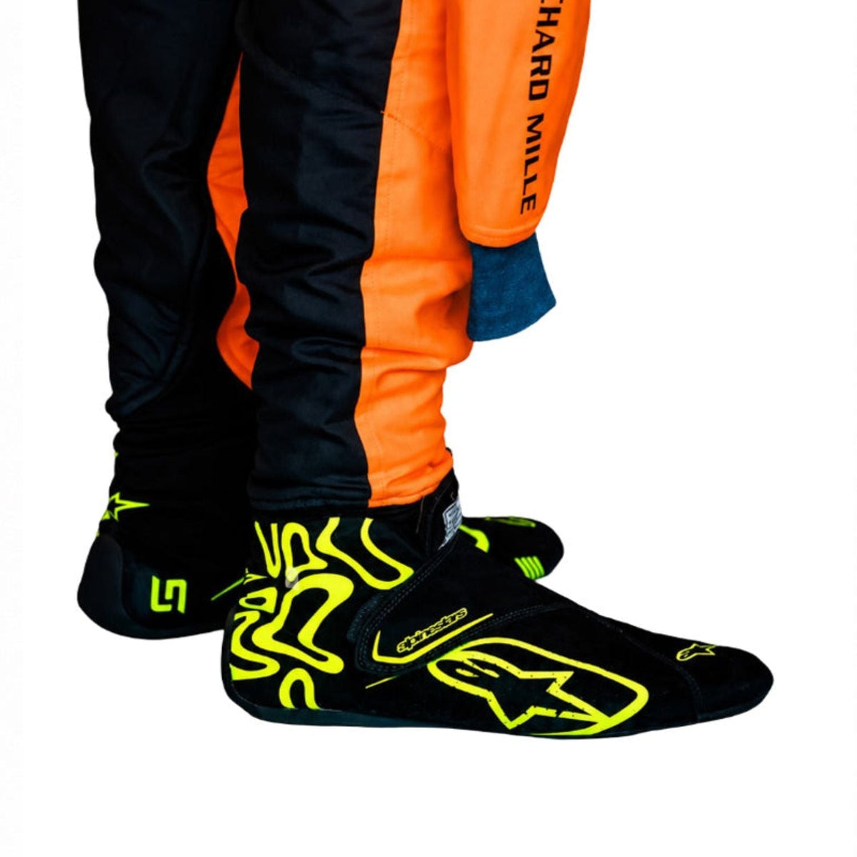 Lando Norris Racing Shoes Replica 2026: close-up of a person's feet wearing Alpinestars racing boots in black with neon yellow graphics, paired with black and orange racing pants.