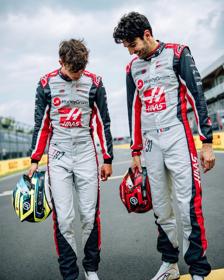 Esteban Ocon Canadian GP 2025 Racing Suit Replica: oliver And Esteban race drivers in matching silver, black, and red Haas F1 racing suits walking on a racetrack, each holding a helmet. Visible branding includes MoneyGram and Haas.
